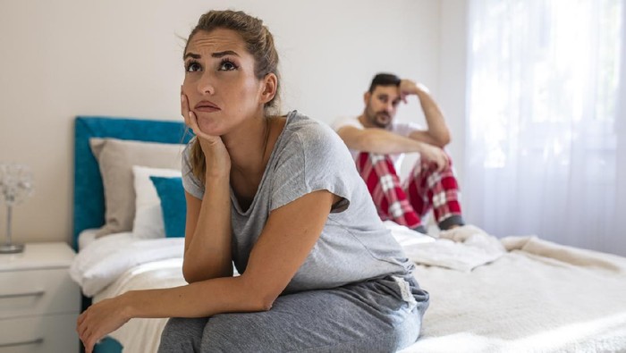 We have a problem. Young upset girl sitting on the edge of the bed, against her boyfriend, lying in bed. Frustrated sad girlfriend sit on bed think of relationship problems, thoughtful couple after quarrel lost in thoughts