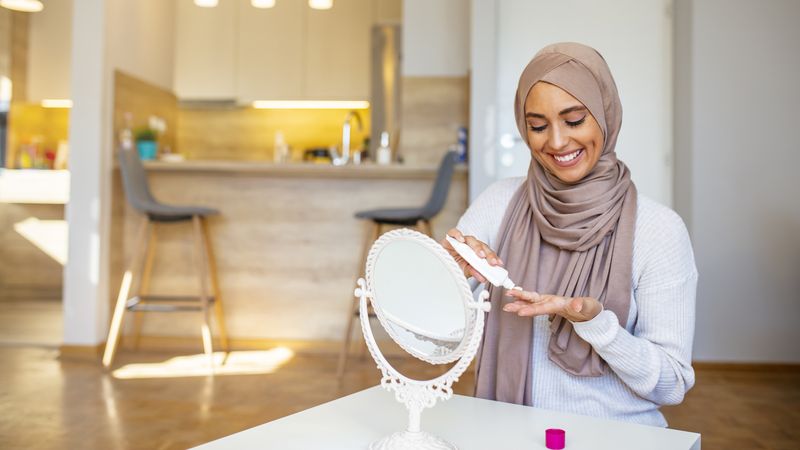 1184700237 Beautiful Muslim woman looking at mirror and putting face cream. Muslim woman with hijab looking at her face in the mirror. Smiling muslim lady applying moisturizing face cream, skin care, cosmetology