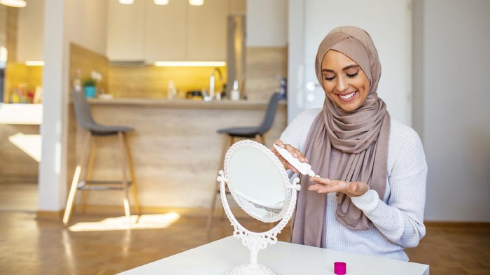 Beautiful Muslim woman looking at mirror and putting face cream. Muslim woman with hijab looking at her face in the mirror. Smiling muslim lady applying moisturizing face cream, skin care, cosmetology
