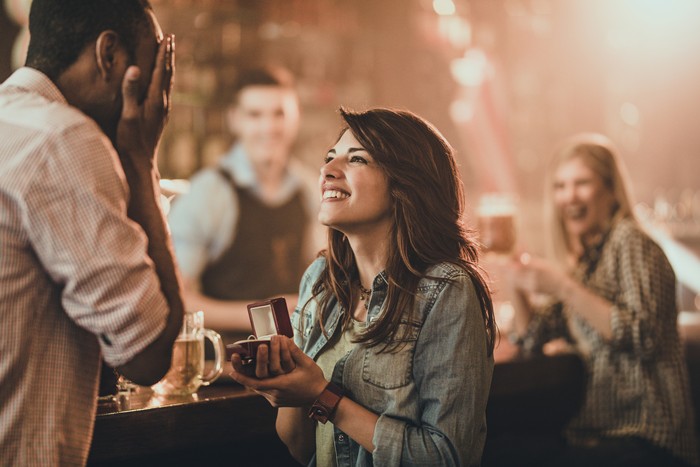 945033420 Young happy woman proposing her black boyfriend during night out in a pub. There are people in the background.