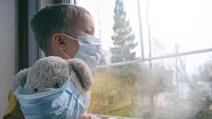 Anak memakai masker Sad illness child on home quarantine. Boy and his teddy bear both in protective medical masks sits on windowsill and looks out window. Virus protection, coronavirus pandemic, prevention epidemic.