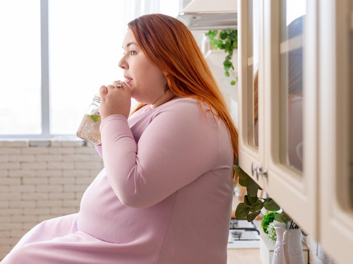 gemuk Relaxation, leisure, rest, domesticity and coziness. Beautiful charming young female with chubby cheeks and curvy body sitting on sofa barefooted, keeping one foot on floor, smiling at camera