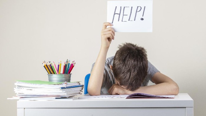 Sad tired frustrated boy sitting at the table with many books and holding help sign. Learning difficulties, education concept.
