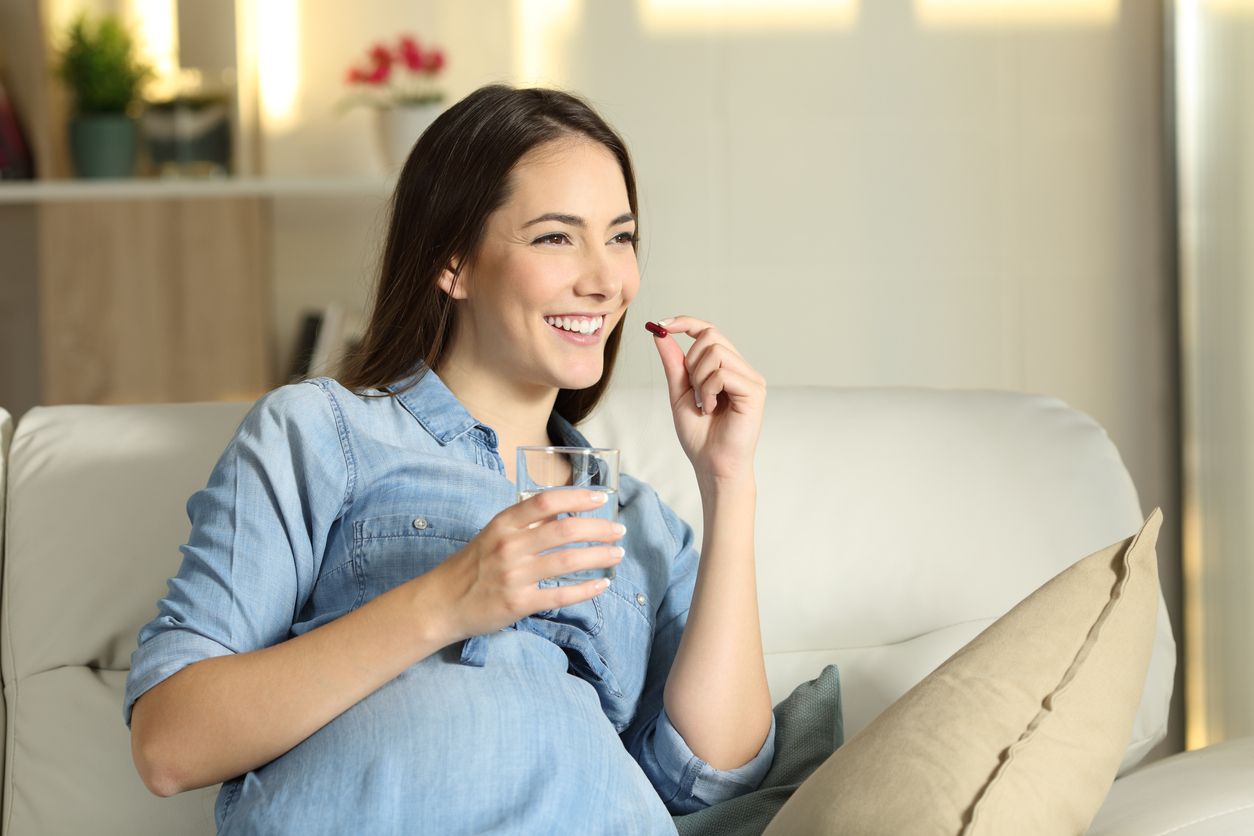 Portrait of a happy pregnant woman taking a pill sitting on a couch in the living room at home