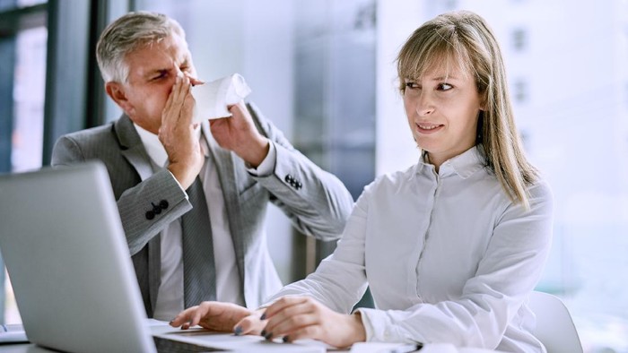 Shot of a sick businessman sneezing in front of a businesswoman in the office