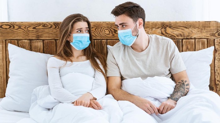 Young male and female in blue protective masks taking photo by phone on tripod at home