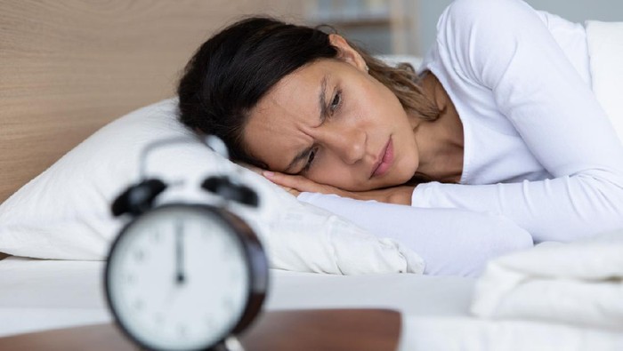 1227225902 Unhappy restless woman wearing pajama suffering from insomnia, alarm clock on bedside table, sleepless tired girl with open eyes lying in bed, lack of sleep, early morning awakening concept