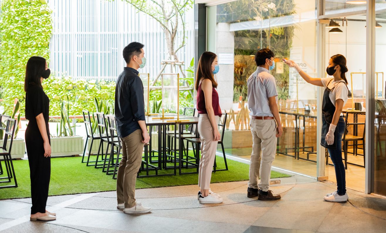 Asian waitress wear protective face mask scan customer temperature by thermometer before entering the restaurant to protect infection from coronavirus covid-19, social distancing concept