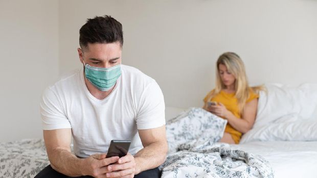 Ilustrasi pasangan Couple under Covid-19 quarantine using their phones at home. Man wearing face mask texting on smartphone.