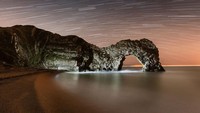 Foto komposit ini menunjukkan pantai yang gelap dan kosong di samping Durdle Door di Pantai Jurassic Dorset, dengan sekitar satu jam rotasi bumi direkam dalam jejak bintang di langit. Jika diperhatikan dengan teliti, Anda mungkin dapat melihat sosok mungil yang sedang menerangi wajah batu dari garis pantai.  