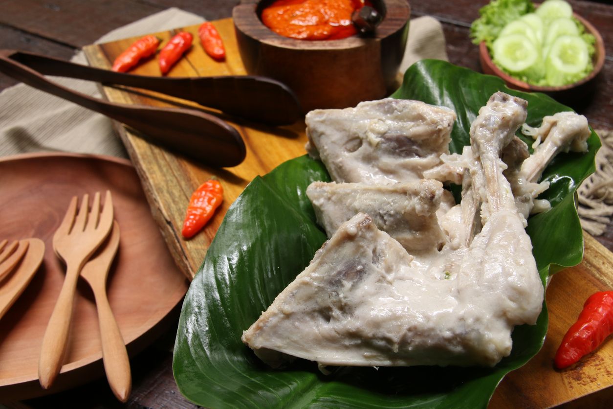 Ayam Pop, a popular chicken dish from Minangkabau, Padang, West Sumatra. The chicken is braised in coconut milk and spices; then only lightly fried. It is served with a special spicy paste of red chili peppers, tomatoes, and broad beans. The chicken pieces are arranged on rectangular wooden serving block that has been lined with a broad green leaf. On the other end is the chili paste. Some chili peppers are scattered on the tray. Some green vegetables of cassava leaves, lettuce and cucumber slices are also served along the chicken dish. A wooden plate with two matching wooden cutleries are placed next to the tray.