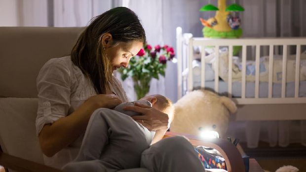 Young beautiful mother, breastfeeding her newborn baby boy at night, dim light. Mom breastfeeding infant