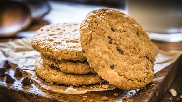 Pile of homemade oatmeal cookies with chocolate chips upon a cutting board on rustic wooden table. Cookies are surrounded by oat flakes, oat spikes and chocolate chips. Main focus is on cookies and in the defocused background is a glass full of milk. Low key DSLR photo taken with Canon EOS 6D Mark II and Canon EF 24-105 mm f/4L