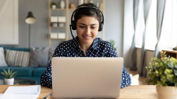 Smiling millennial Indian girl in wireless headset sit at desk in living room study online on laptop, happy young ethnic female in headphones watch webinar or course on Internet on computer