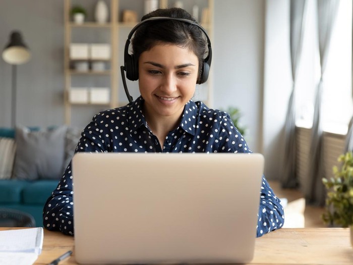 Smiling millennial Indian girl in wireless headset sit at desk in living room study online on laptop, happy young ethnic female in headphones watch webinar or course on Internet on computer