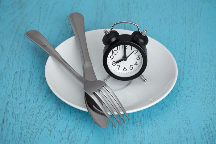Ilustrasi diet Intermittent fasting concept with clock on white plate, fork and knife on blue table