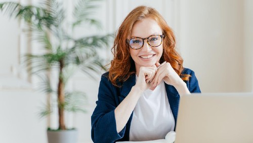 Young Caucasian busy brunette smiling and holding pencil while sitting in office and looking at laptop.