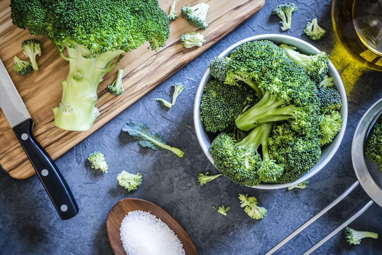 Top view of a bowl full of broccoli branches surrounded by a wooden cutting board with a entire broccoli and a kitchen knife on top, an olive oil bottle, a metal colander and a little wooden tray with salt on a grey textured backdrop. Low key DSLR photo taken with Canon EOS 6D Mark II and Canon EF 24-105 mm f/4L