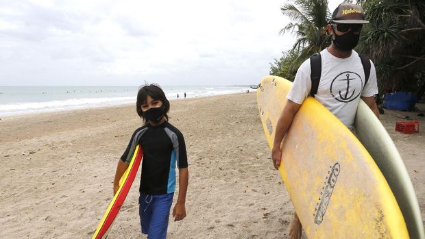Tourists wearing face masks carry their surfboard at Kuta beach, Bali, Indonesia on Thursday, July 9, 2020. Indonesia's resort island of Bali reopened after a three-month virus lockdown Thursday, allowing local people and stranded foreign tourists to resume public activities before foreign arrivals resume in September.(AP Photo/Firdia Lisnawati)