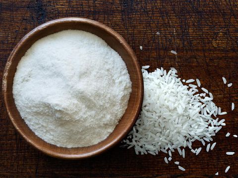 White rice flour in dark wooden bowl isolated on dark brown wood from above. Spilled rice.