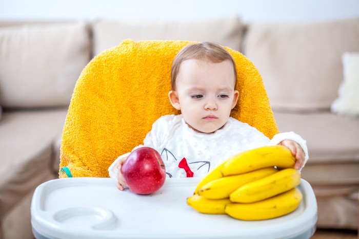 Little baby girl sitting in baby chair eating fruit apple and bananas