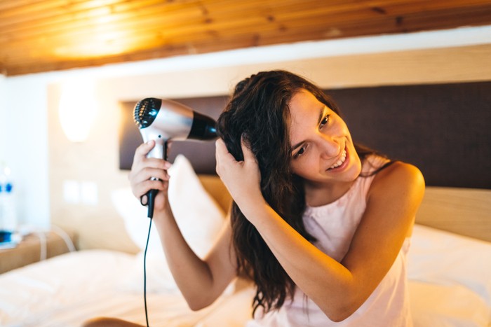 Young smiling brunette, sitting on the bed, drying her hair. Home interior. Waist up.
