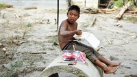 Seorang anak tampak bersantai makan di sebuah pipa beton sambil bertelanjang dada.