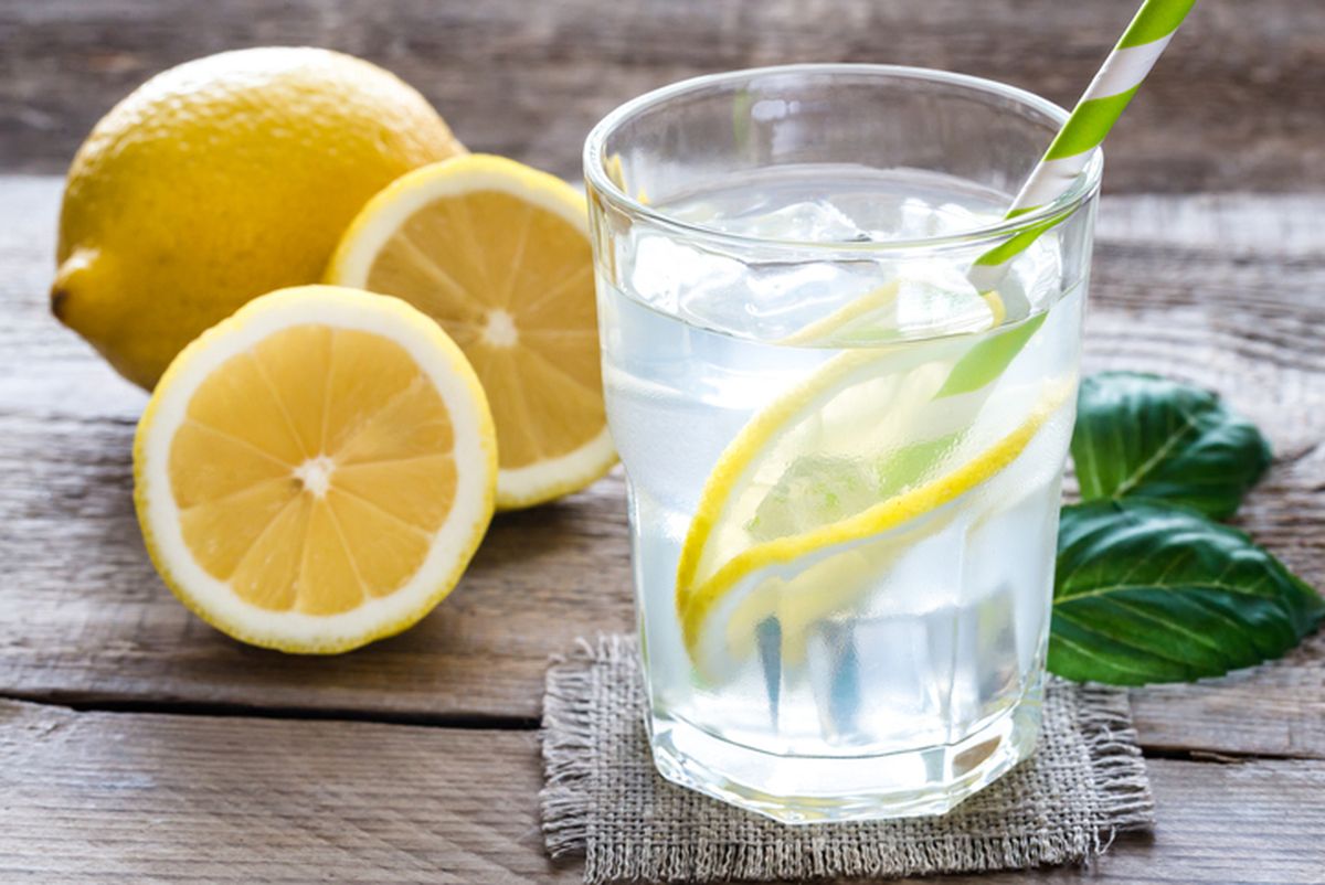 Segelas air lemon segar berada di atas meja dengan latar belakang taman hijau, menampilkan irisan lemon dan dedaunan mint yang menambahkan sentuhan segar