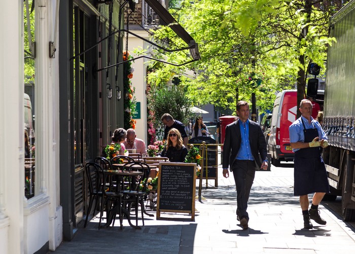 London, UK - 21 May, 2019: color image depicting people sitting at tables, eating and drinking outside a posh cake shop/cafe in the Belgravia area of London, UK. The exterior of the shop is decorated with beautiful garlands of fresh flowers. It is a beautiful day with bright blue sky. Room for copy space.