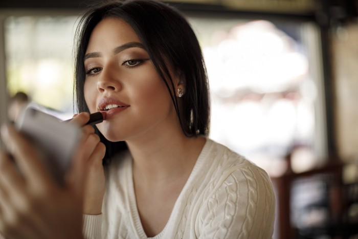 1137392664 Young woman applying lipstick at a cafe