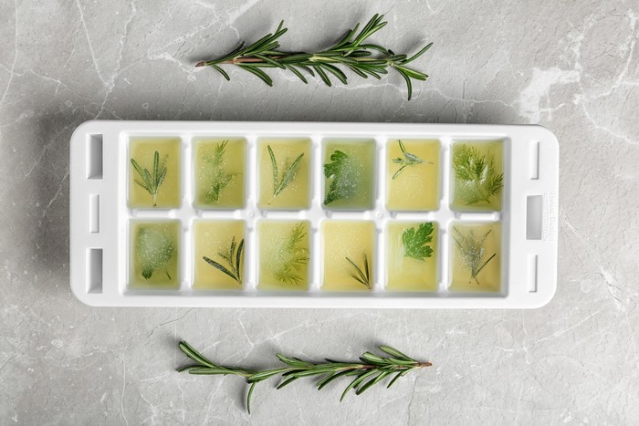 Ice cube tray with herbs frozen in oil and fresh rosemary on grey background, flat lay