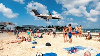 Turis-turis berdiri di sekitar tepi pantai, menanti pesawat lewat dan merekam momen-momen burung besi itu melintas di atas kepala dengan ponsel atau kamera. (iStock)