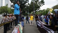 Pemimpin aksi berorasi dengan berdiri di atas pembatas jalan. Sementara peserta aksi lainnya membentangkan spanduk dan poster.