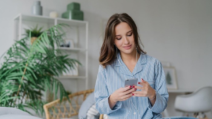 Photo of relaxed young European lady in pyjamas enjoys audio playlist, listens music romantic songs in earphones, uses modern cell phone, sits on comfortable bed against domestic atmosphere.