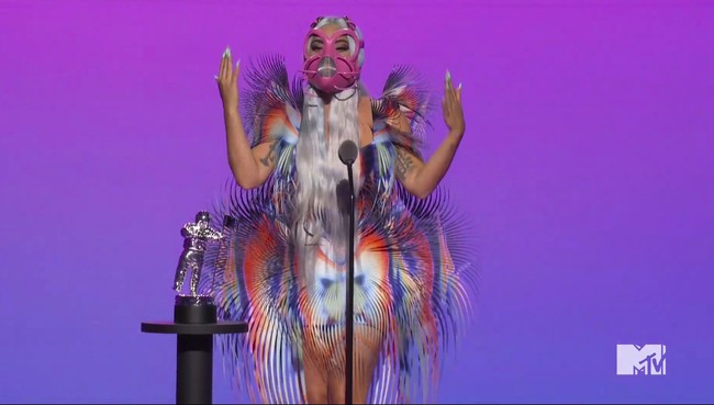 Di MTV VMA 2020, Lady Gaga mendapat sembilan nominasi (terbanyak dari nomine lainnya). Best Collaboration untuk lagu Rain on Me, buah kolaborasinya dengan Ariana Grande, menjadi penghargaan pertama yang disabetnya. Untuk momen tersebut, ia tampil nyentrik dalam balutan gaun Iris van Herpen dengan rumbai warna-warni, berikut masker pink yang hampir menutup seluruh wajahnya. (Foto: MTV via AP)