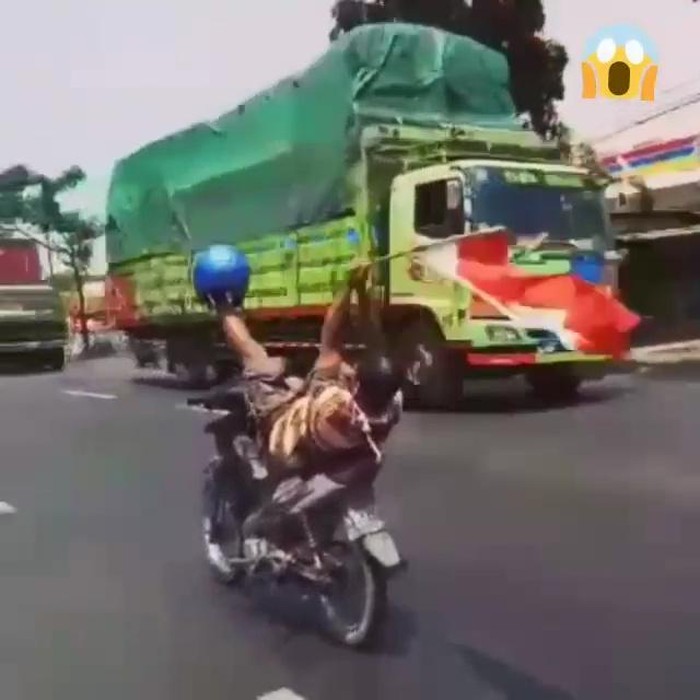 Seorang pemotor nekat mengendalikan setir dengan satu kaki di jalan raya. Sambil tidur di atas jok, pria tersebut mengibarkan Bendera Merah Putih.