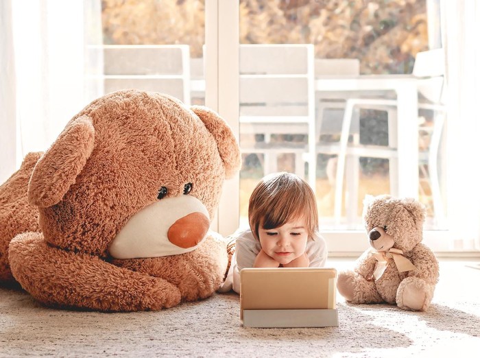 Cute little child watching cartoons on digital tablet device lying on floor with two soft teddy bear toys at home. Modern childhood.
