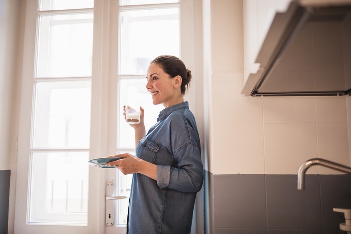 Ilustrasi wanita minum susu Young woman eating a croissant, drinking fresh milk for breakfast in the kitchen. She is wearing only a blue shirt, standing by the big, bright window