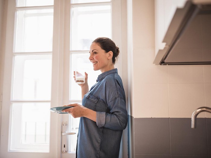 Young woman eating a croissant, drinking fresh milk for breakfast in the kitchen. She is wearing only a blue shirt, standing by the big, bright window