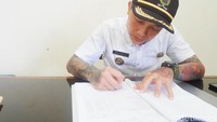 Ditemui detikcom hari ini, Hoho terlihat tengah sibuk di ruang kerjanya. Ia memakai kemeja putih lengan pendek lengkap dengan topi Kades. Di bagian lengan nampak tato dengan motif oriental.