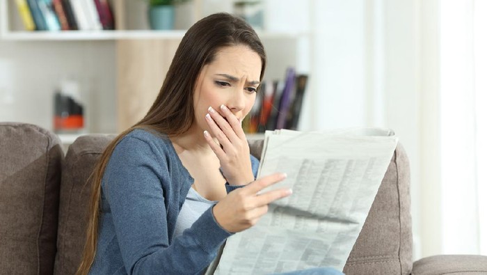 Worried woman reading bad news in a newspaper sitting on a couch in the living room at home