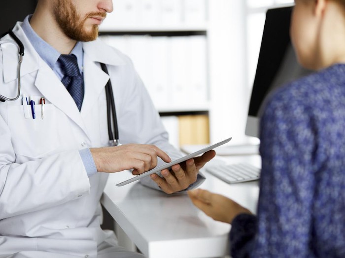 1254405995 Unknown red-bearded doctor and patient woman discussing current health examination while sitting and using tablet computer in clinic, close-up. Medicine concept.