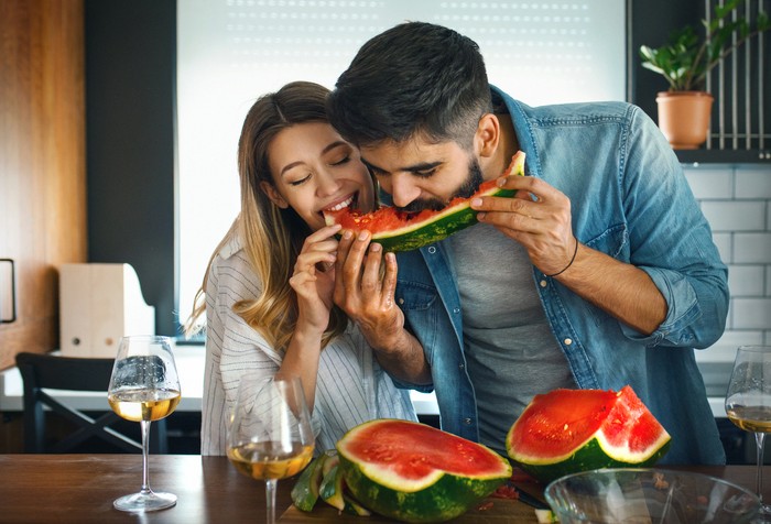 Closeup front view of a mid 20s couple sharing a watermelon in a kitchen. They are biting the same slice and laughing, having some white wine as well