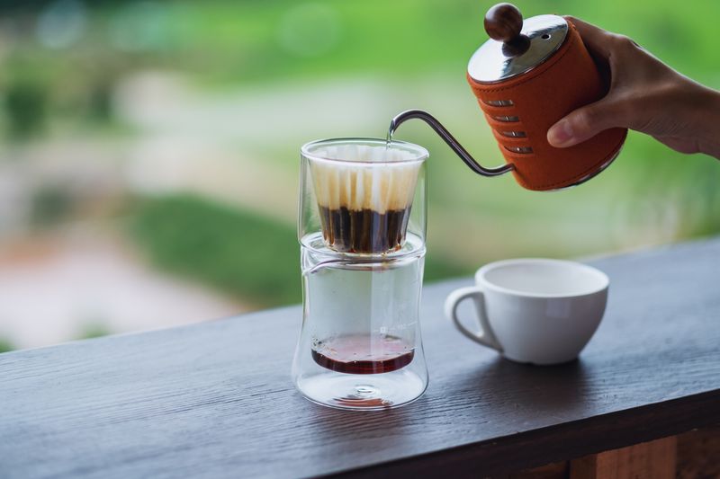 Closeup image of a hand pouring hot water from kettle to make drip coffee with blurred nature background