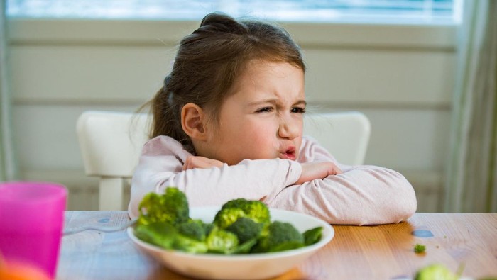 Cute girl eating spinach and broccoli at the table. Child doesnt want to eat, refuses eating, making faces. Healthy food concept.