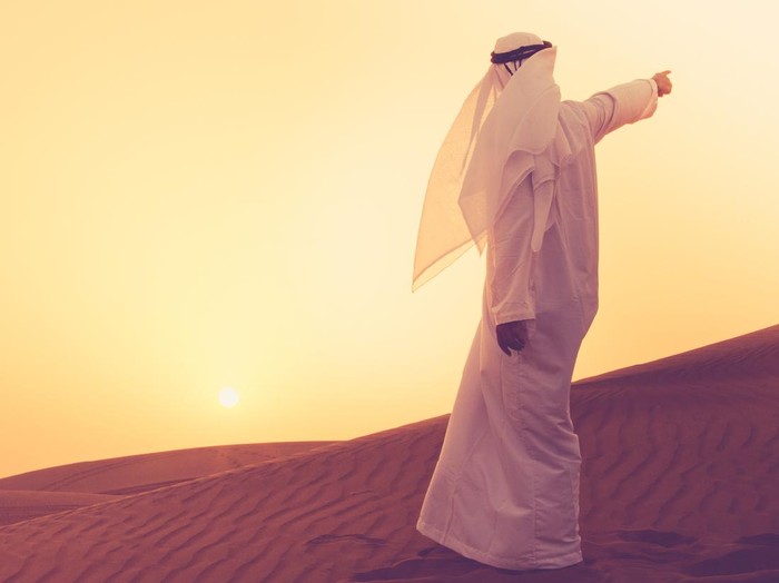 An Arab standing at the sand dunes of Dubai and aiming towards the horizon.