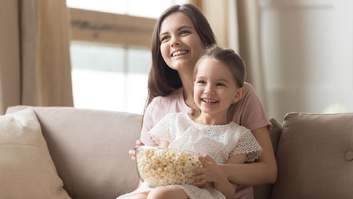 menonton film Happy young mom and child girl laughing holding snack popcorn remote control enjoy funny television comedy movie, smiling family mother with kid girl watching fun humor tv show laugh sitting on sofa
