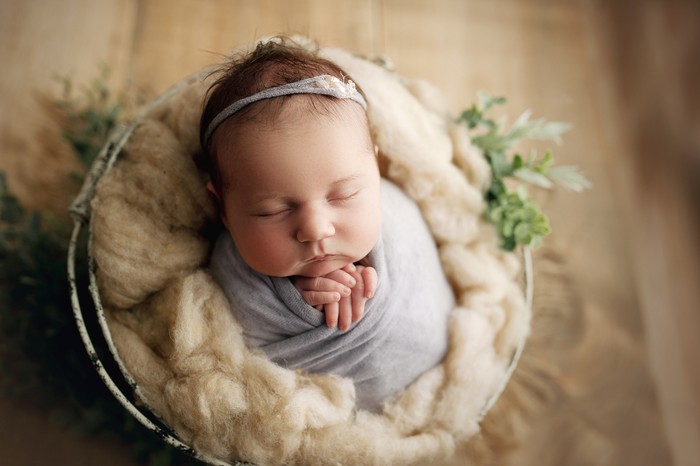 20 day old baby girl with chunky cheeks, swaddled in pale purple with a matching headband, in a prop basket for her newborn milestone photos