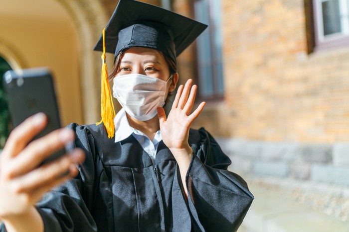 A student in graduation gown is making a video call with a smart phone.