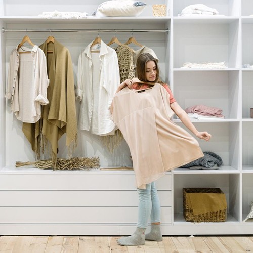 Young adult woman standing near modern wardrobe at home with contemporary interior, choosing trendy clothes, having difficult choice not knowing what to put on, trying on fashionable wear